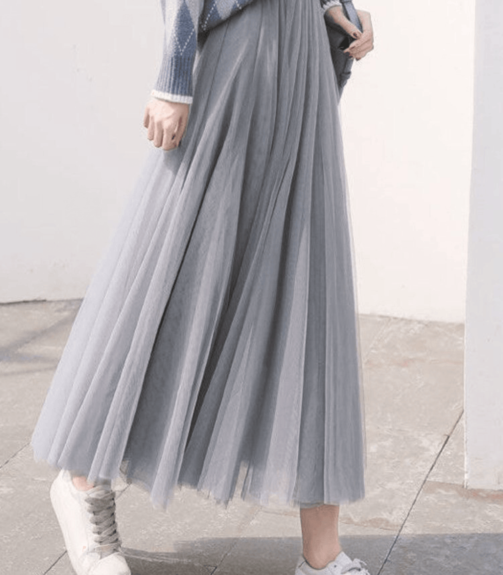 Vue de face d’une femme portant une jupe longue plissée grise à taille haute avec un haut gris clair et des chaussures blanches.