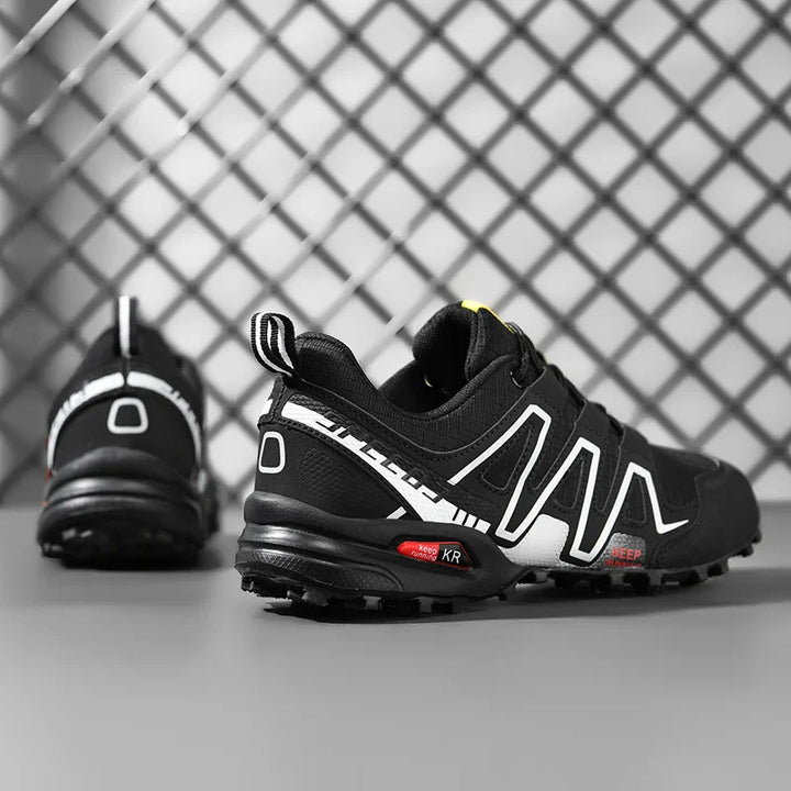 Chaussures trail noires avec lignes contrastées, vue de côté devant un grillage, focus sur l’avant et l’adhérence de la semelle
